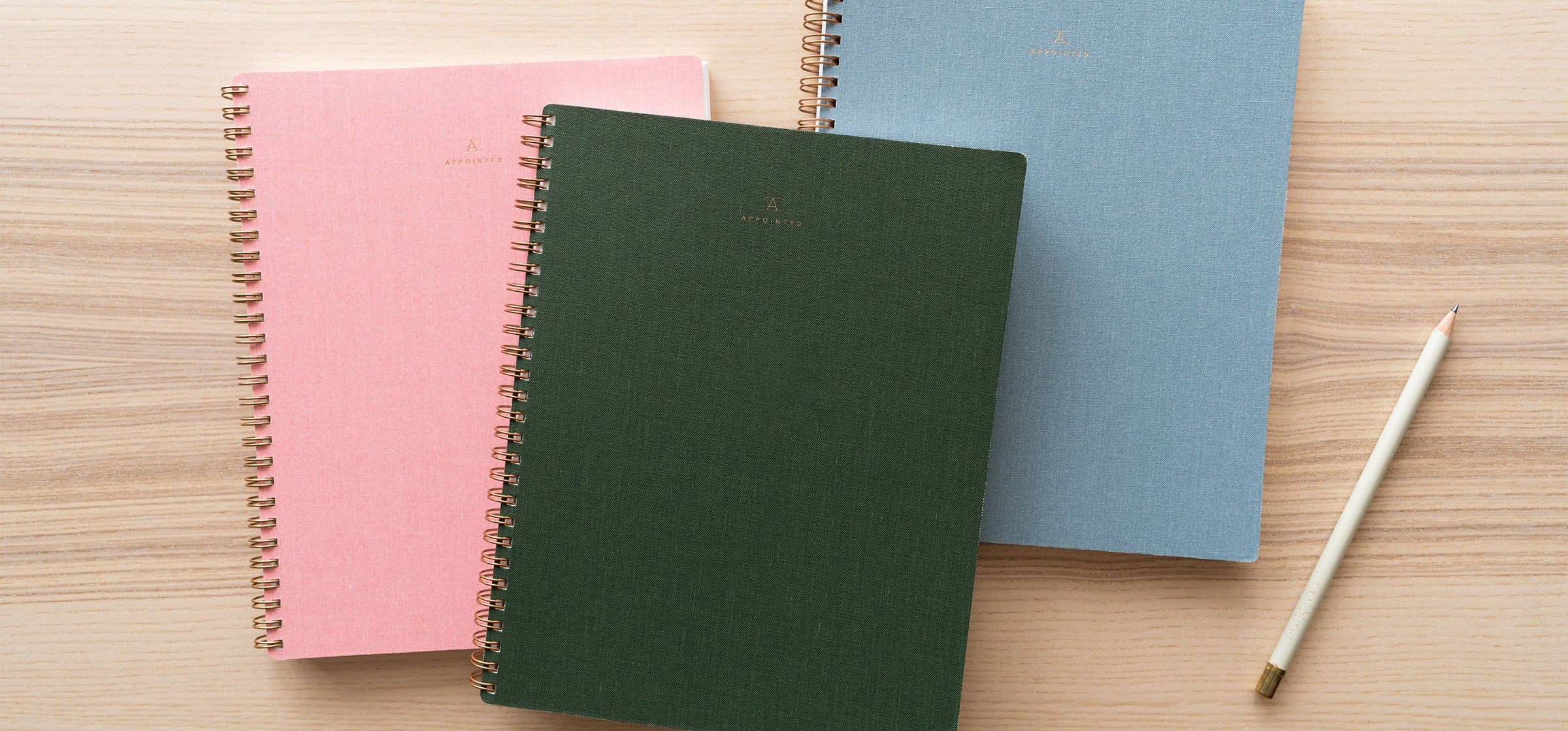 Blossom Pink, Hunter Green, and Chambray Blue Notebooks sit on top of a wood desk with a no. 2 pencil aside them