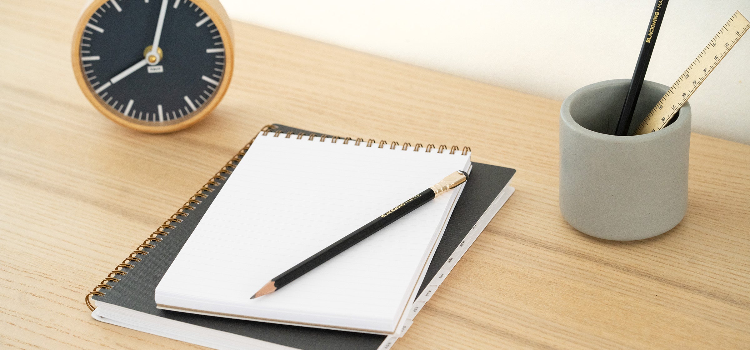 A serene desk scene shows an analog clock, stack of paper goods, and writing tools in a ceramic vessel.