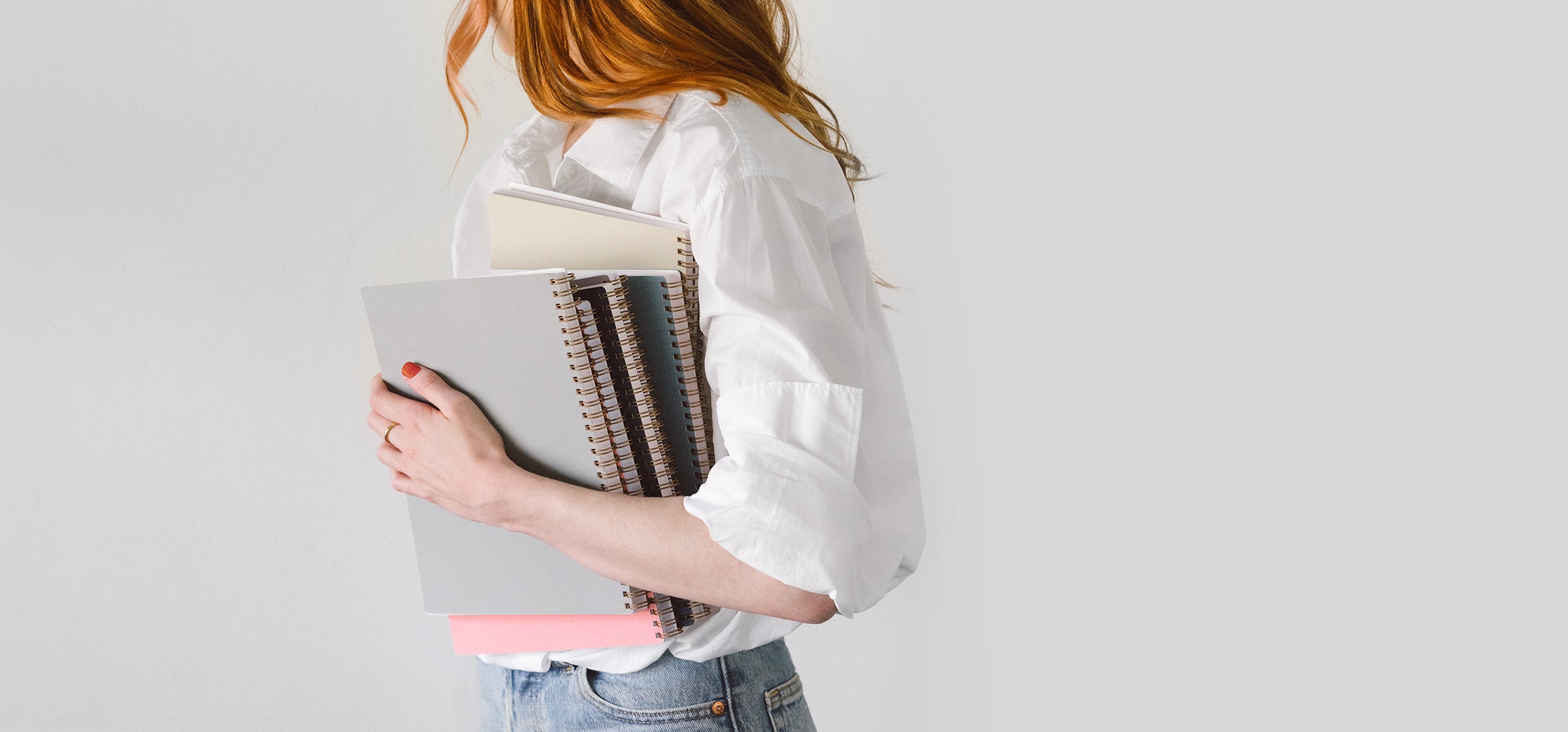 A woman with long red hair wears an oversize white button-down and jeans. she stands against a gray wall holding a stack of Appointed notebooks in her left hand while she looks away from the camera.